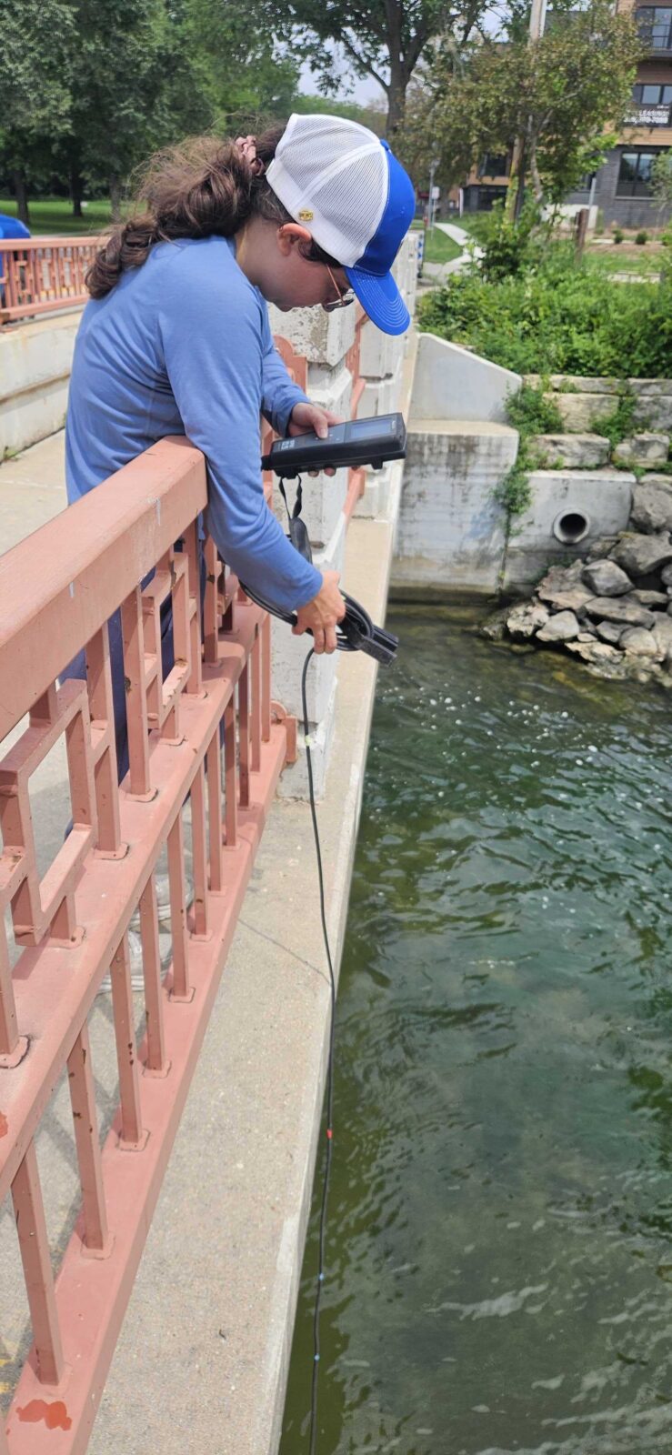 A person lowers a temperature and dissolved oxygen sensor into a river from a bridge and reads the screen.