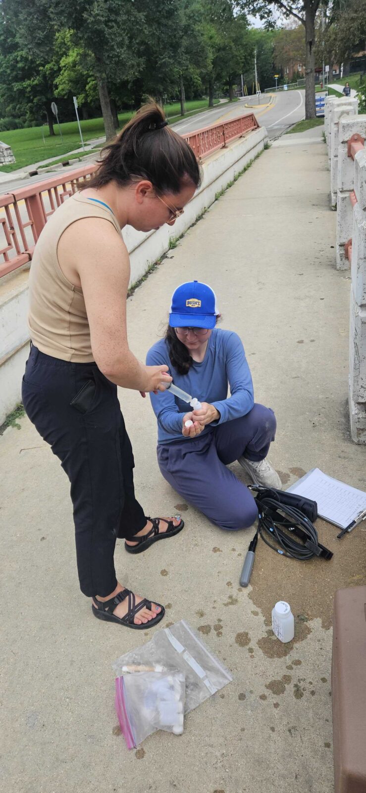 One person filters water into a vial being held by a second person at a stream sampling site.