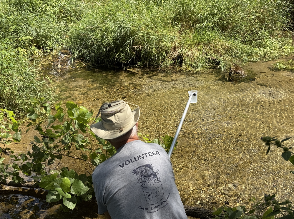 A person takes a water sample from a headwater spring using an outstretched sampling device.